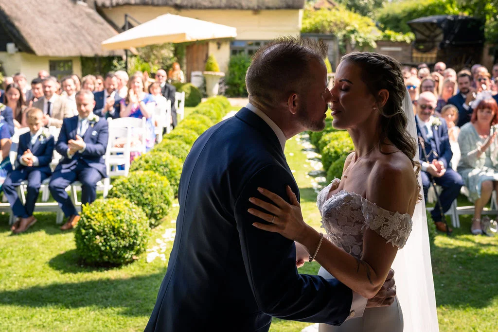 A newlywed couple share their first kiss during an outdoor garden wedding ceremony surrounded by seated guests on a sunny day.