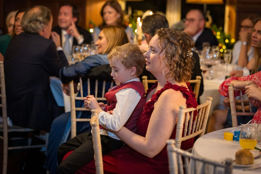 A young page boy in a red polka dot waistcoat sits comfortably on the lap of a guest wearing a red dress during a wedding reception.