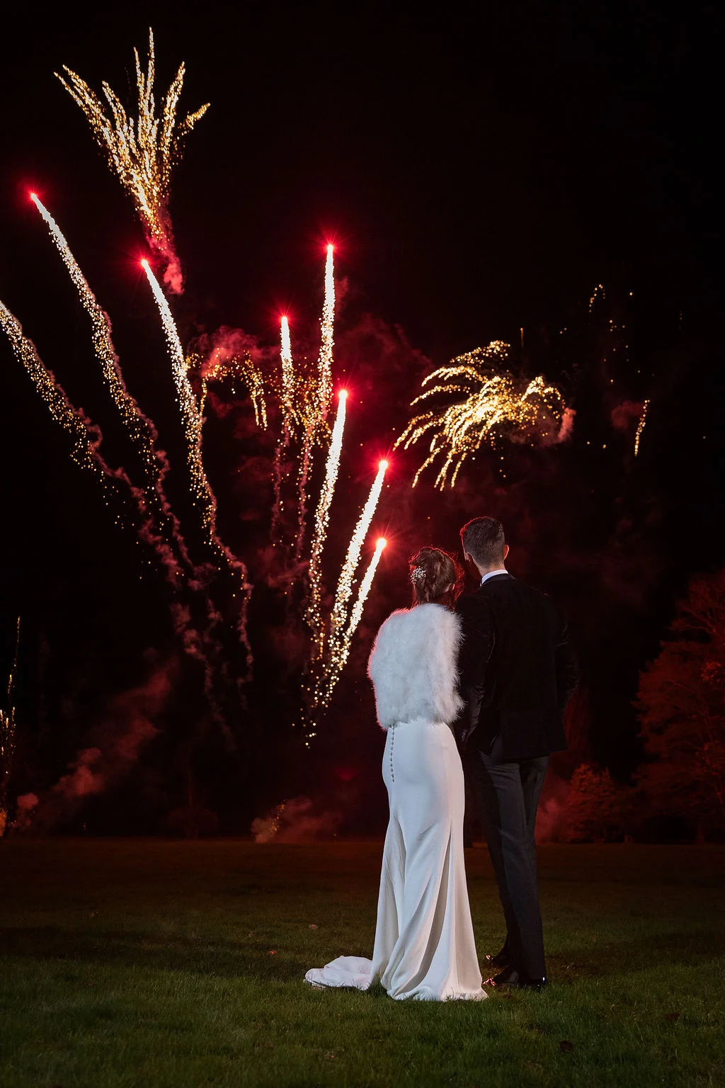 Newlyweds stand outdoors at night, viewed from behind, watching a spectacular fireworks display light up the dark sky.