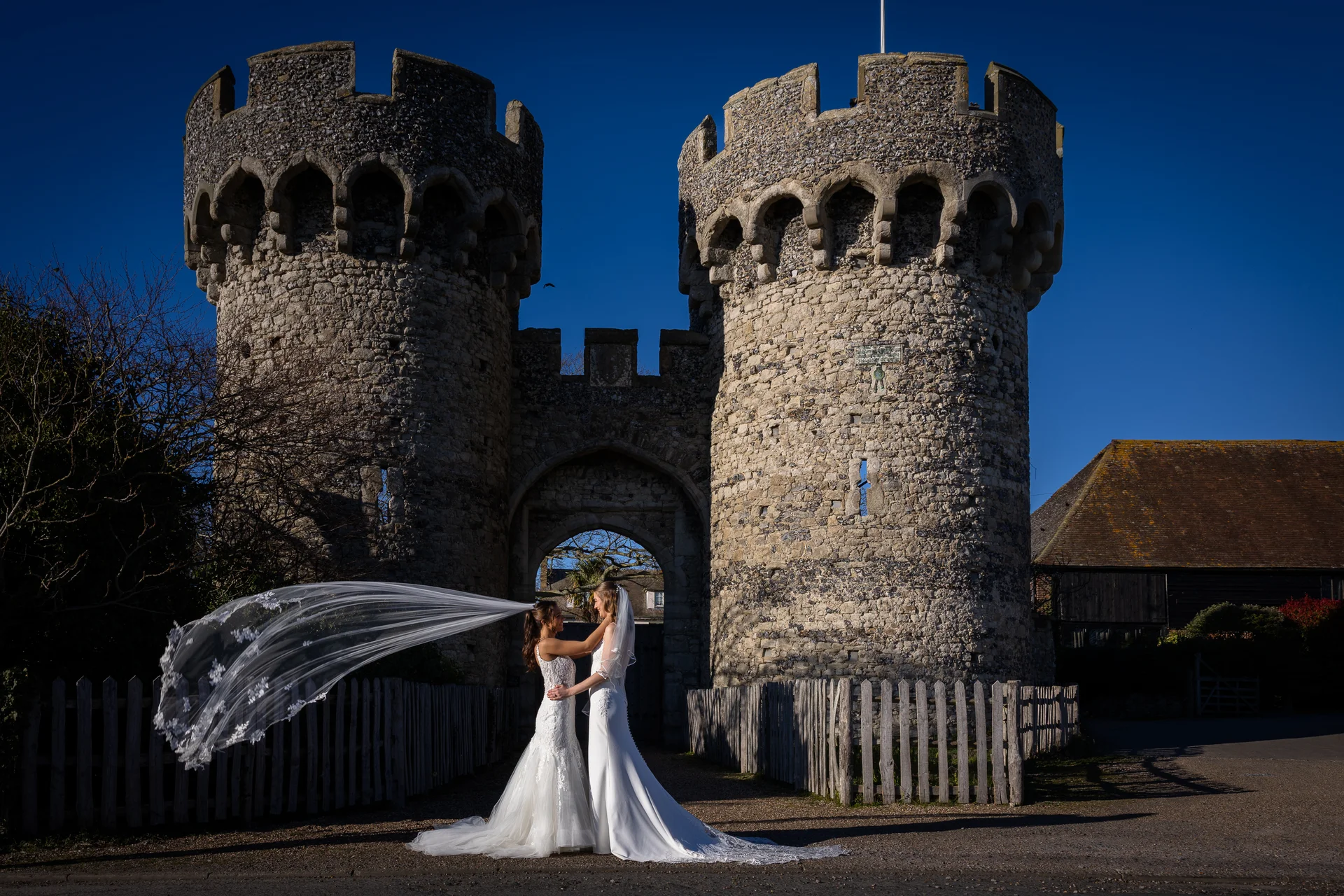 Two brides in stunning white wedding gowns embrace in front of a historic stone castle gatehouse as a long lace-edged veil blows dramatically in the wind.