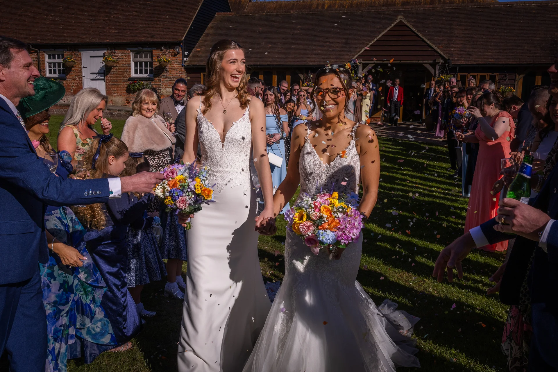 Two smiling brides holding hands and carrying colourful floral bouquets walk through a joyful shower of confetti thrown by guests outside a sunny rustic barn venue.