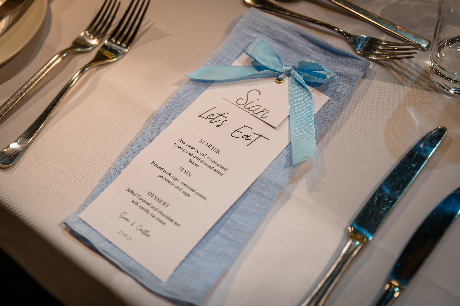 An elegantly styled wedding place setting features a pale blue linen napkin tied with a matching ribbon and topped with a personalised menu card.