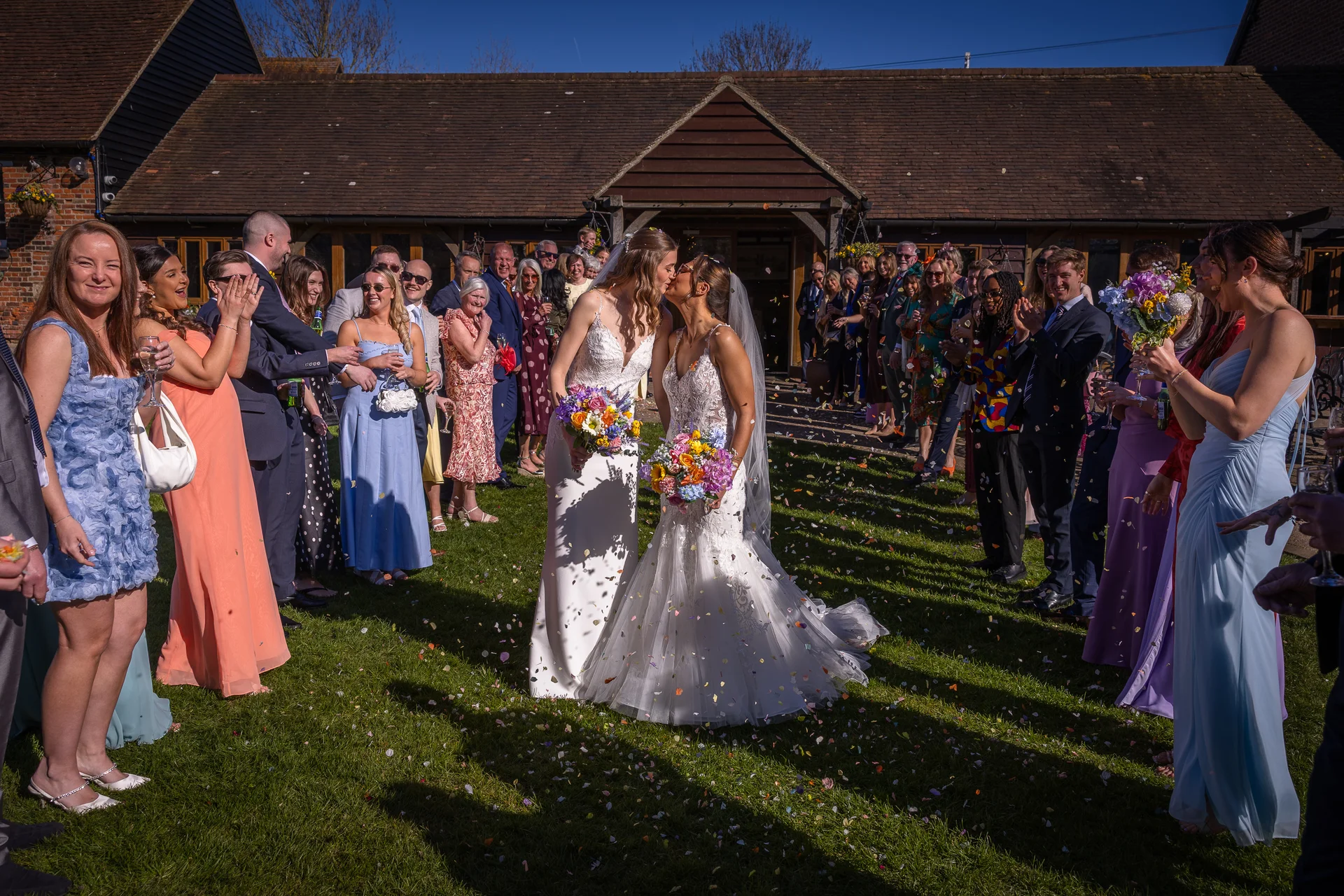 Two brides share a kiss on the lawn outside a rustic barn as wedding guests cheer and shower them with colourful confetti during a sunny summer reception.