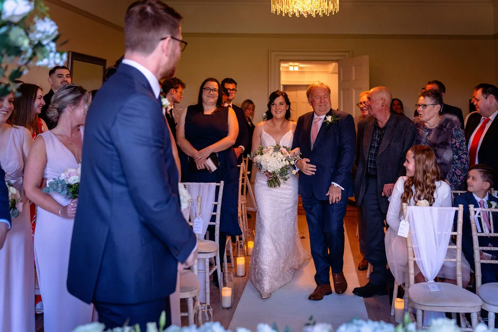 The bride walks down the aisle with her father, smiling towards the groom as guests look on during the indoor wedding ceremony.