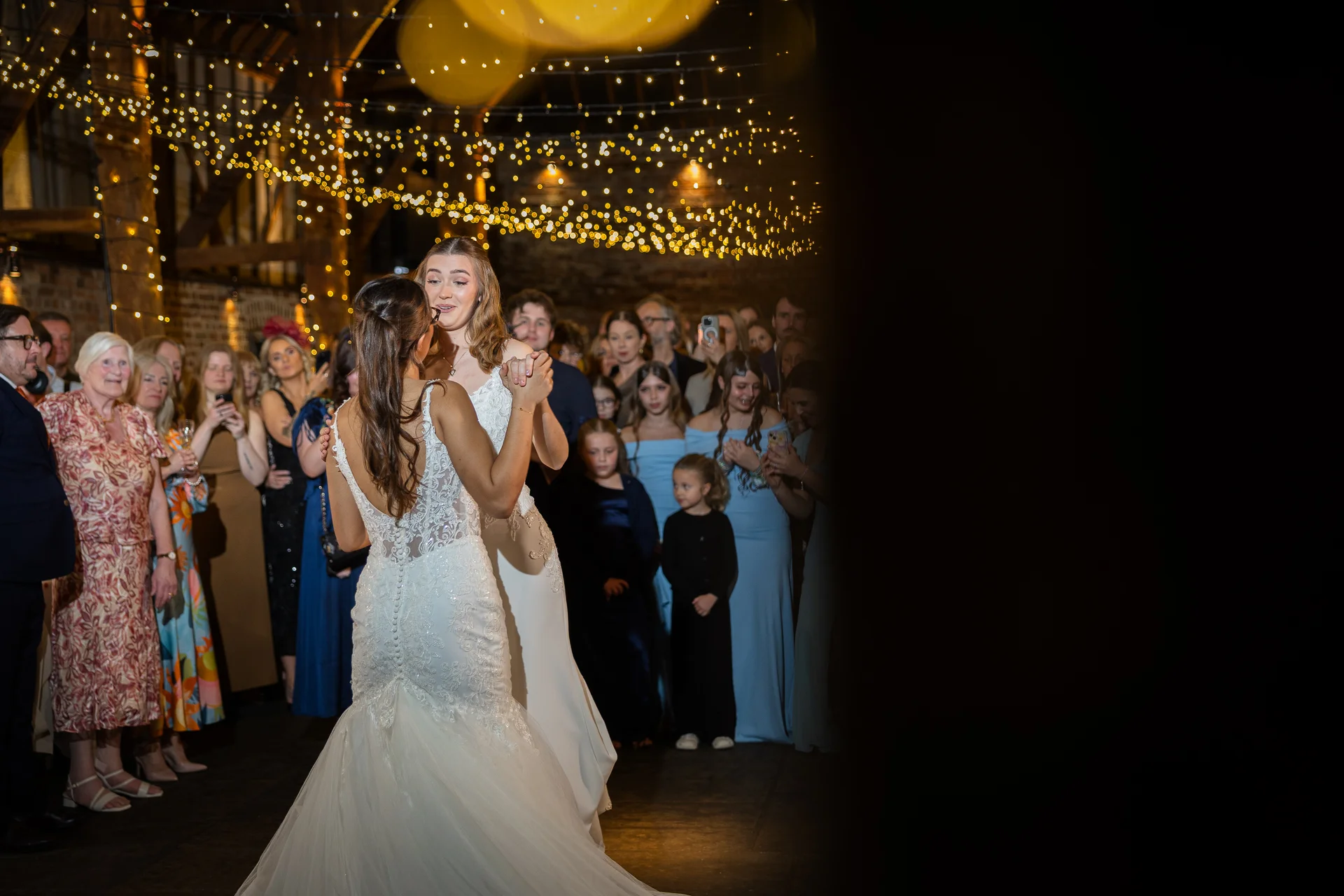 Two brides share their first dance together under a glowing canopy of fairy lights inside a rustic barn while wedding guests look on.