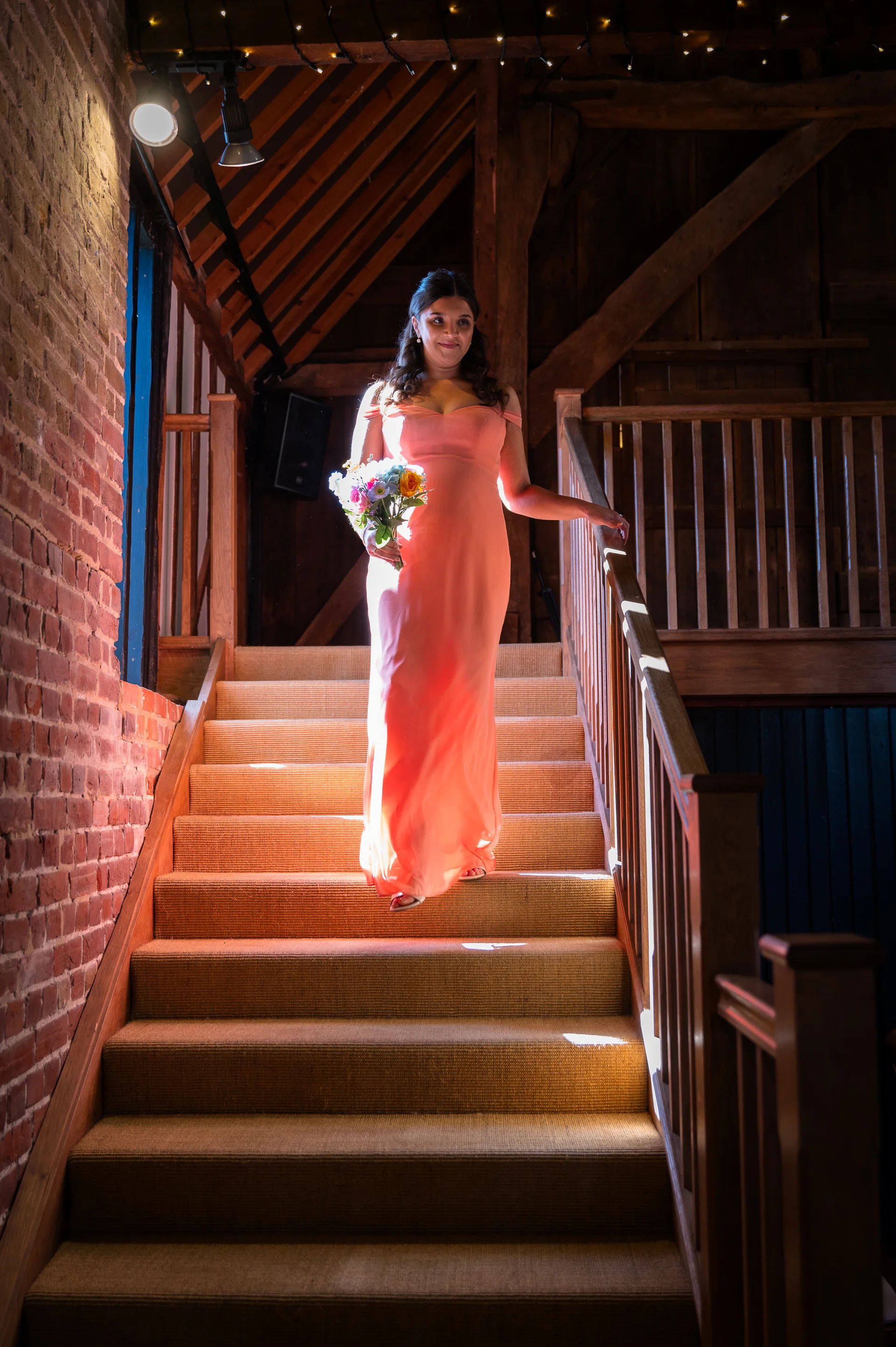 A smiling bride wearing a coral off-the-shoulder gown walks down a wooden staircase holding a colourful bouquet inside a rustic barn.