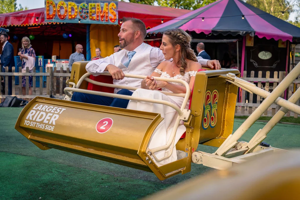 A bride and groom enjoy a vintage funfair ride together during their festival themed wedding reception, surrounded by colourful fairground attractions.