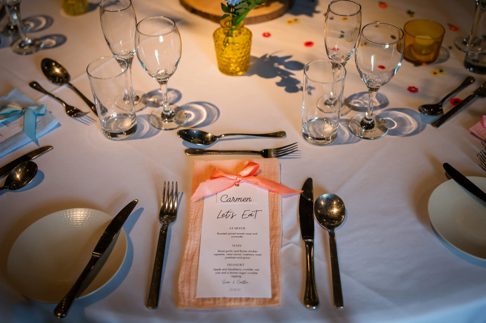A beautifully laid wedding tablescape features white linen, polished cutlery, crystal glassware, and a printed menu card tied with a peach ribbon on a peach napkin.