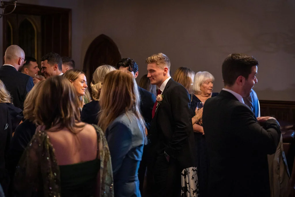 A candid photograph capturing guests mingling and chatting indoors during a wedding reception in a dimly lit hall.