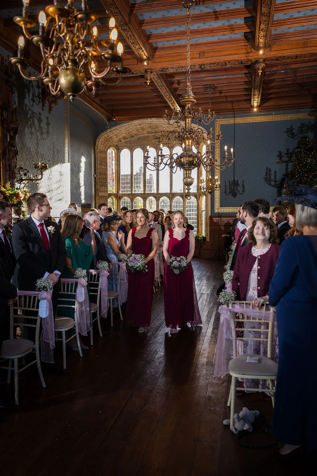 Bridesmaids in burgundy dresses walk down the aisle during a ceremony inside a grand castle venue featuring wooden floors and ornate chandeliers.