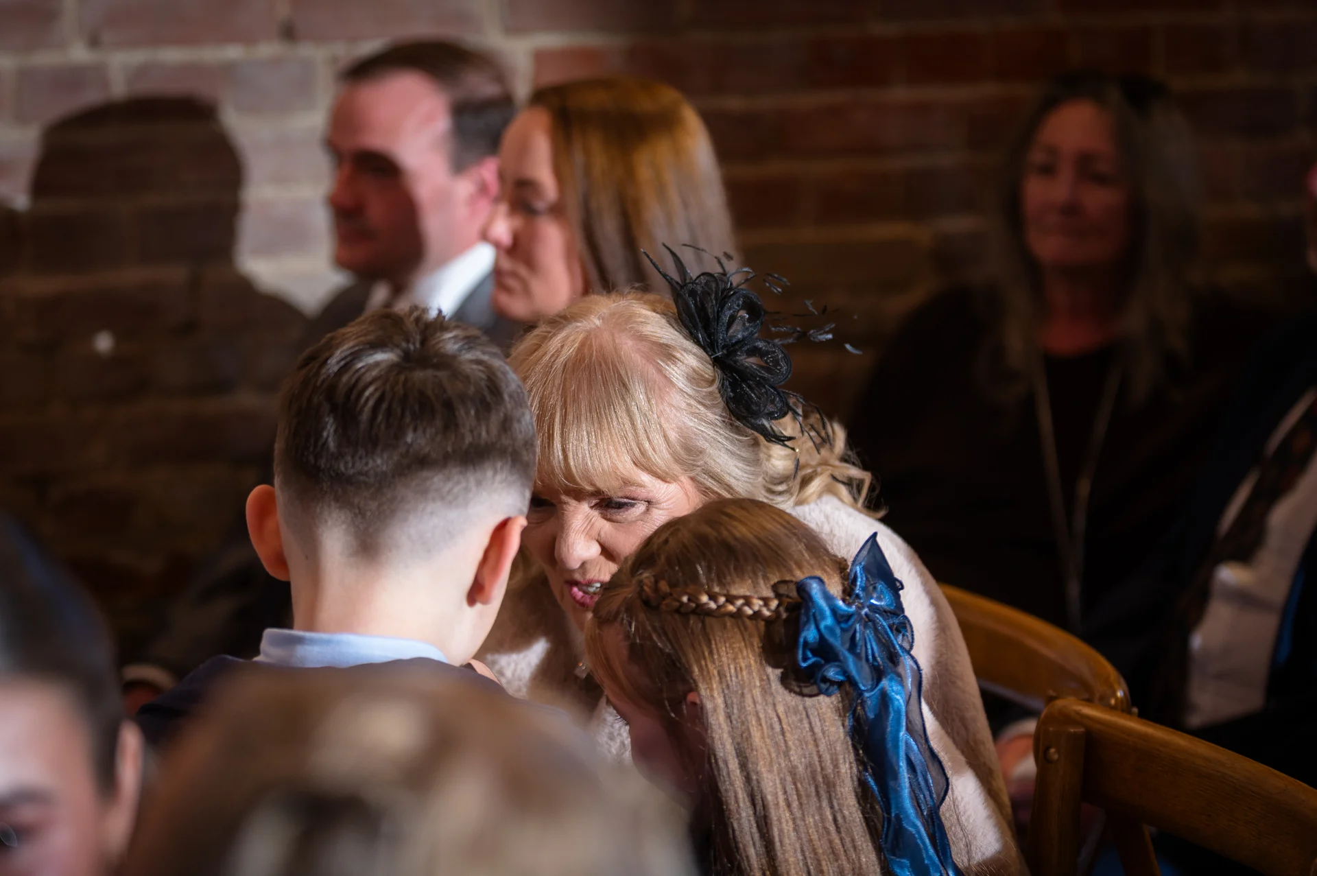 An older woman wearing a black fascinator shares a tender moment with children at a wedding ceremony seated in a venue with exposed brick walls.