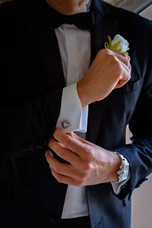 A groom dressed in a sharp black tuxedo adjusts his cufflinks while preparing for the wedding ceremony.