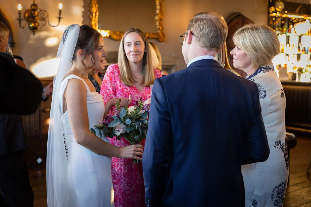 The bride holding a bouquet and veil chats with guests during a lively drinks reception inside a historic venue.
