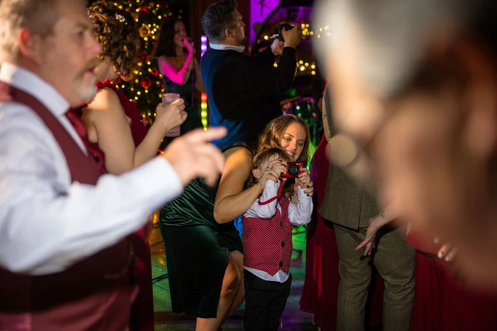 A young child holding a small camera while adults celebrate on a lively dance floor illuminated by festive fairy lights.