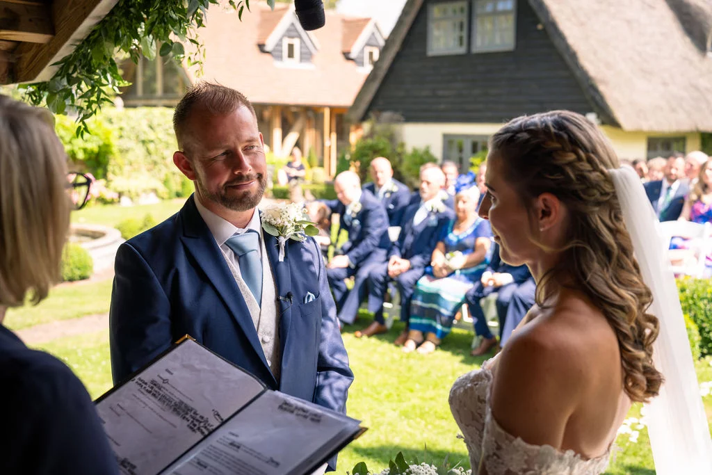 A smiling groom in a navy suit looks lovingly at his bride during an outdoor garden ceremony under bright sunshine.