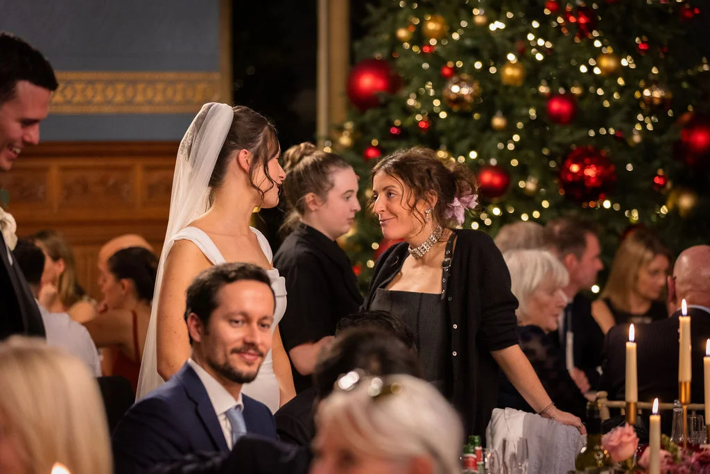 A bride in a white gown and veil shares a warm moment during a festive wedding reception illuminated by glowing Christmas lights and candles.