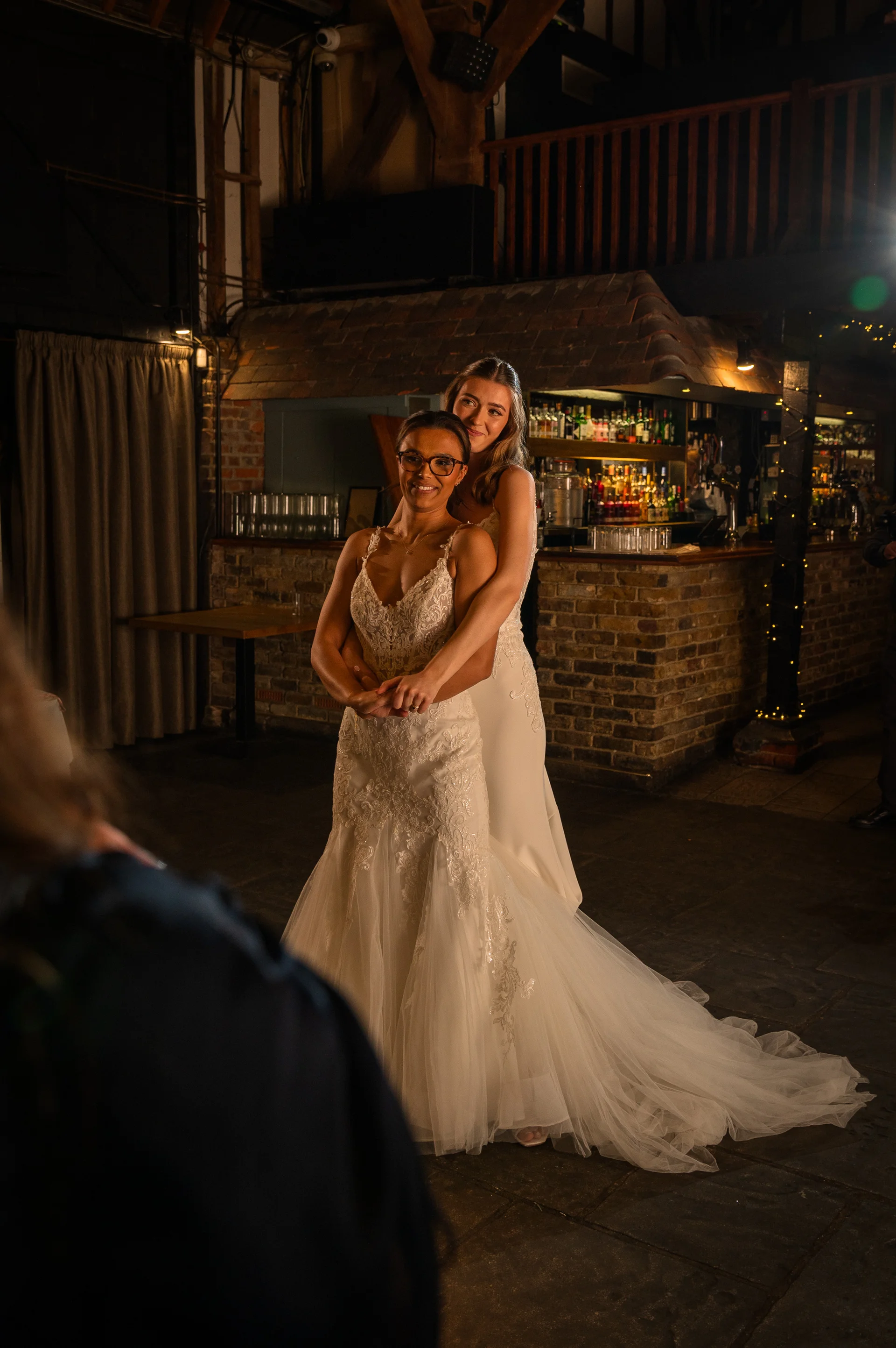 Two brides pose lovingly together inside a rustic barn venue at night, with warm lighting illuminating their wedding gowns and the brick bar backdrop.