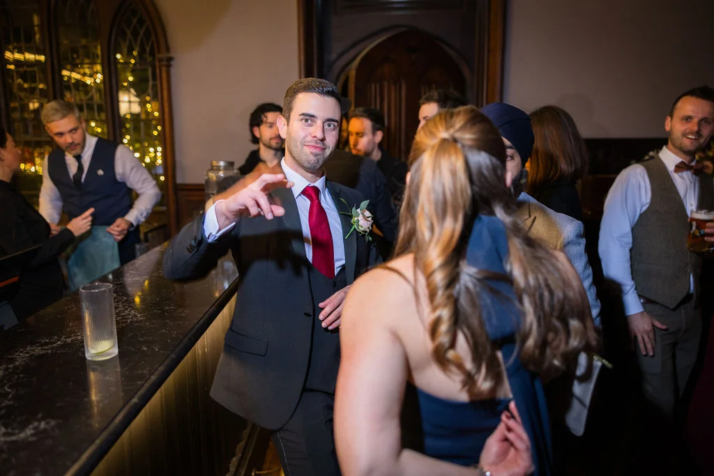 A groom in a dark three-piece suit with a red tie gestures cheerfully while chatting with guests at a lively pub wedding reception.