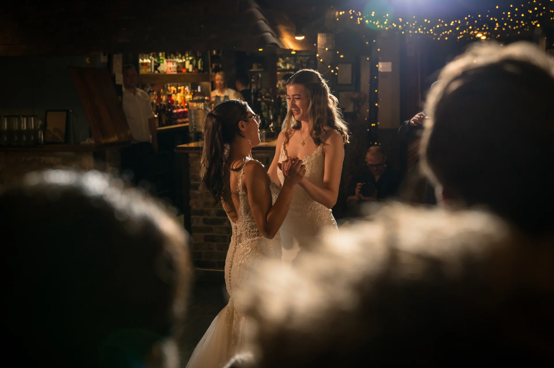 Two brides share a tender first dance together in a cosy pub venue illuminated by warm fairy lights.