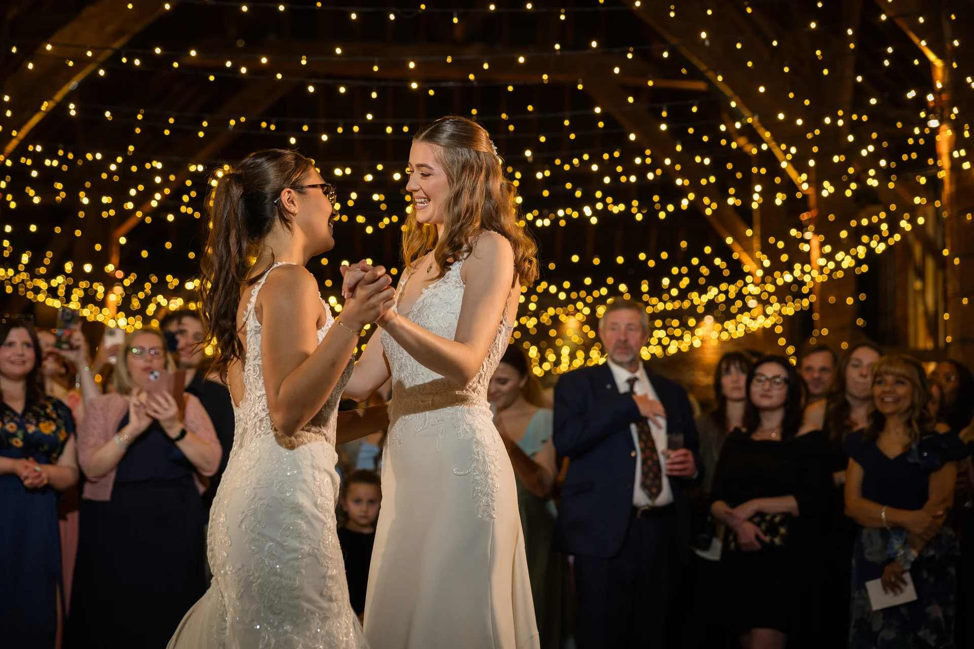 Two smiling brides share their first dance in a rustic barn illuminated by a stunning canopy of warm fairy lights while guests watch on.