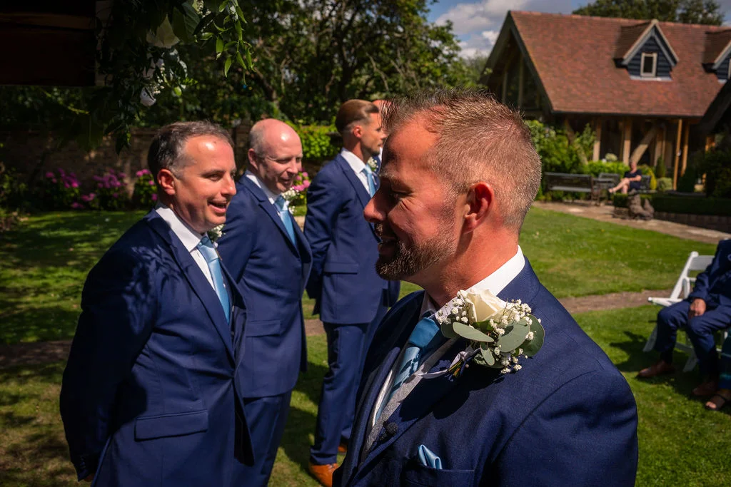 The groom and his groomsmen wearing blue suits stand smiling in a sunny garden setting during an outdoor wedding ceremony.