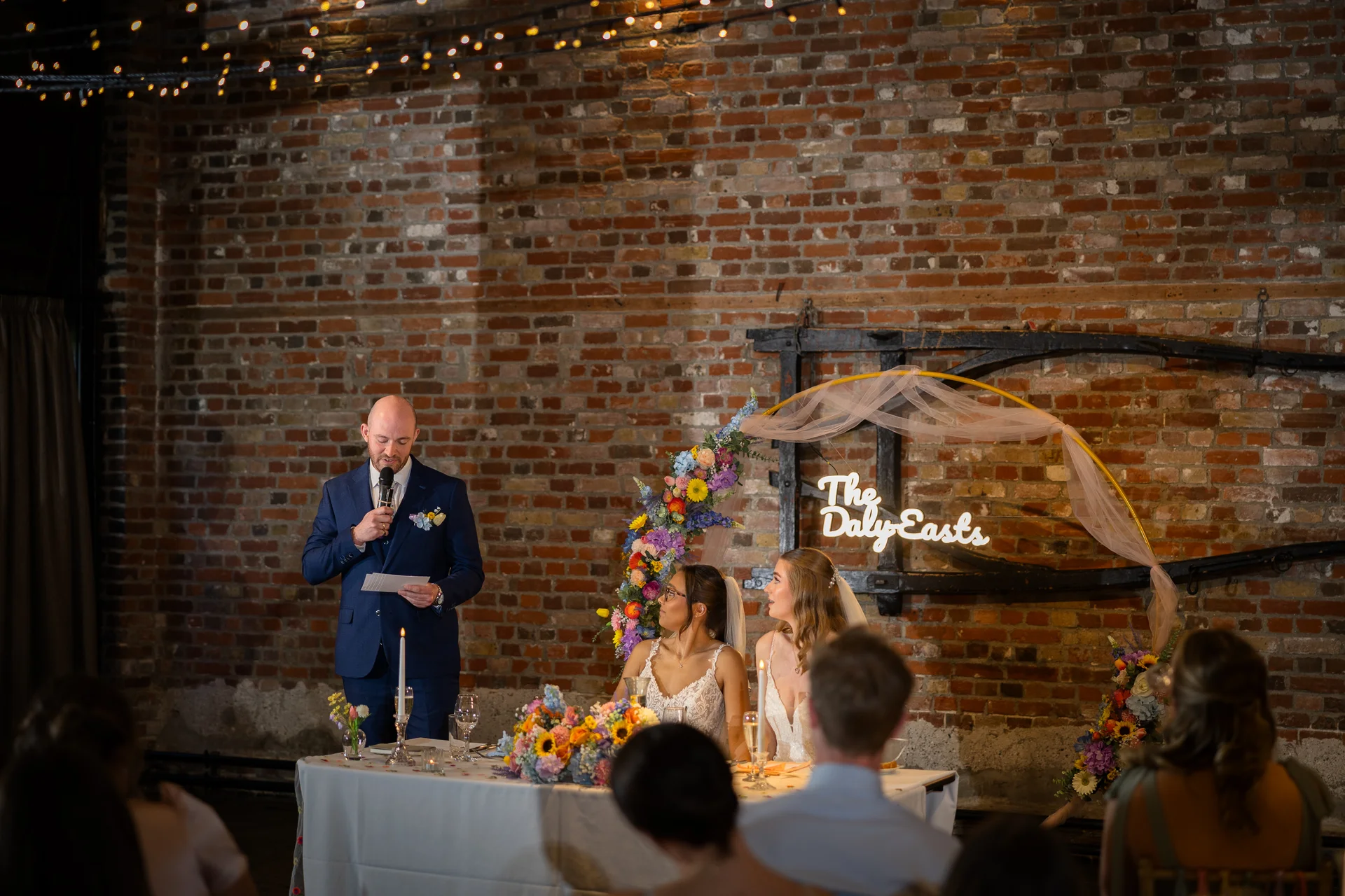 A guest delivers a speech with a microphone at the top table during a warehouse wedding reception in front of an exposed brick wall and neon sign.