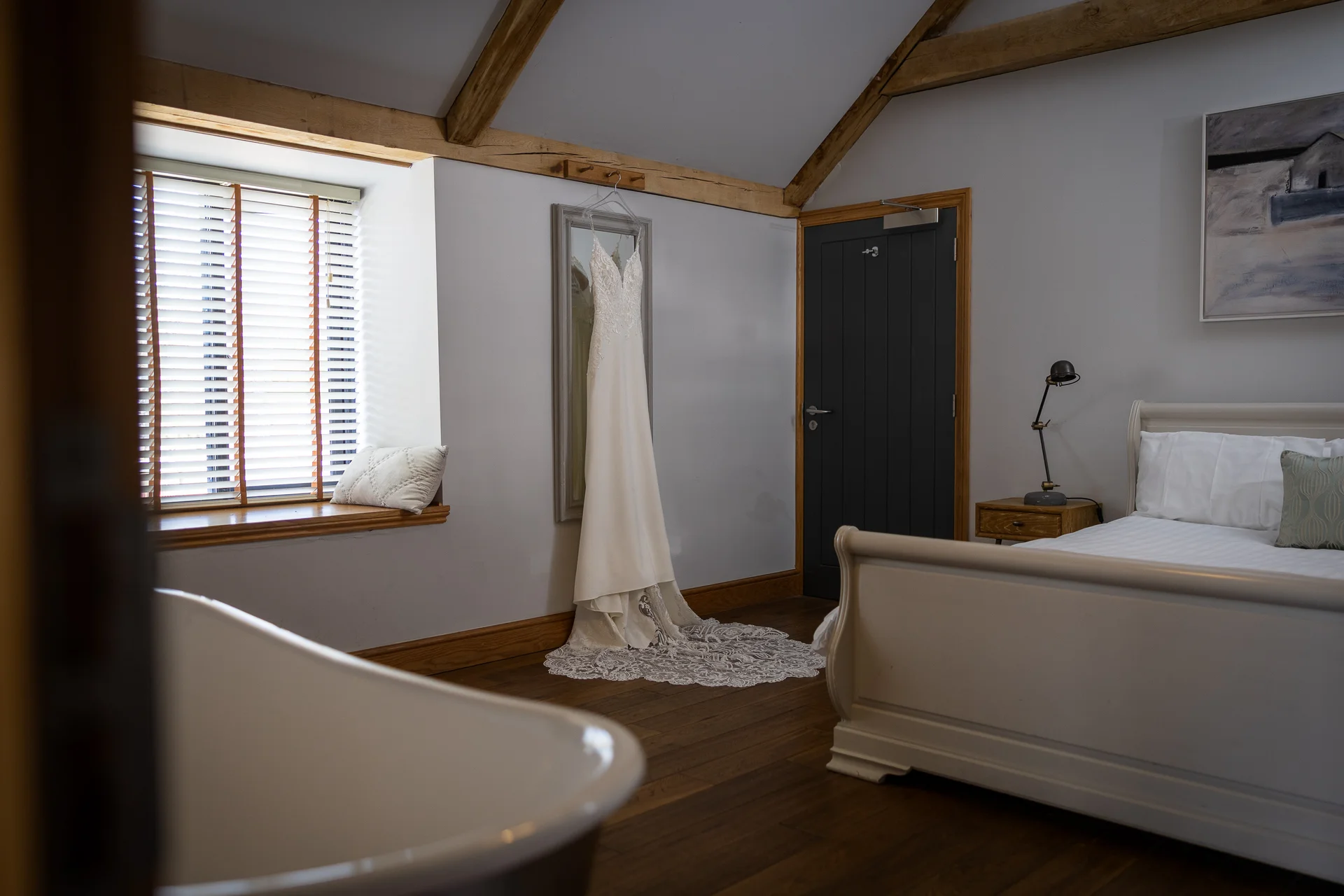 An elegant wedding dress hangs gracefully against a grey wall in a bright bridal suite featuring rustic wooden beams and a freestanding bathtub in the foreground.
