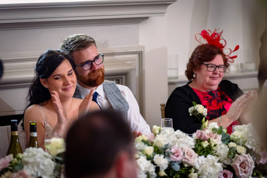 A smiling bride and groom sit at the top table clapping during wedding speeches, accompanied by a guest wearing a red fascinator and floral arrangements.
