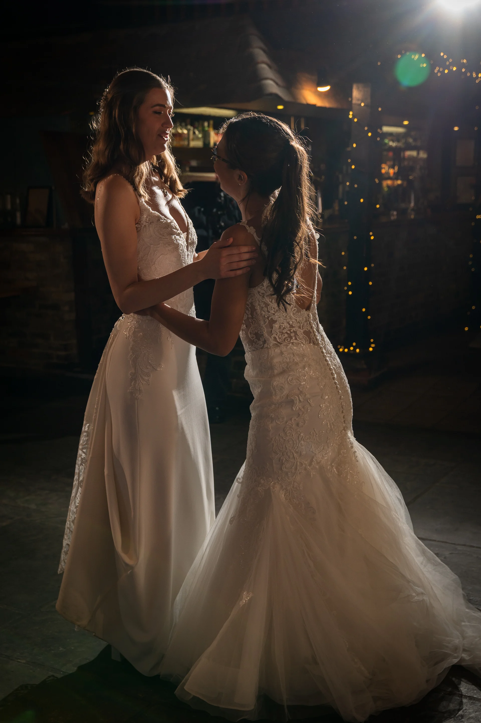 Two brides share a tender moment during their first dance inside a dimly lit venue, beautifully silhouetted against background fairy lights.