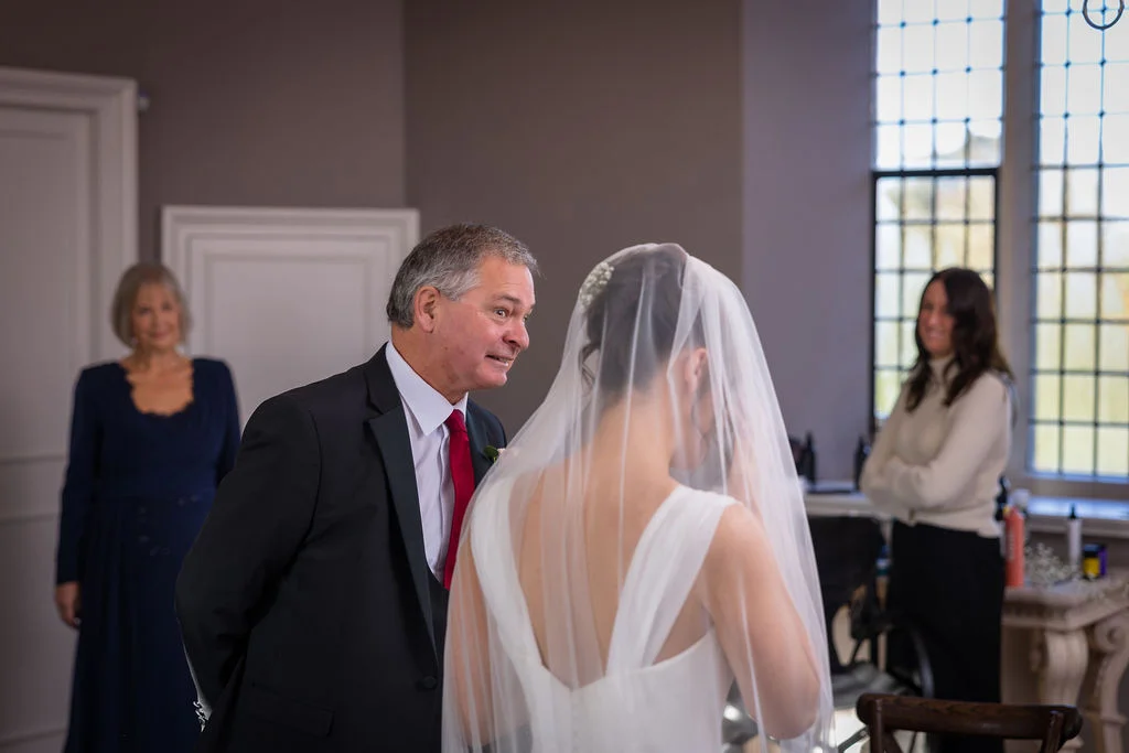 An emotional father of the bride wearing a suit and red tie looks proudly at his daughter in her wedding dress with a veil during morning preparations.