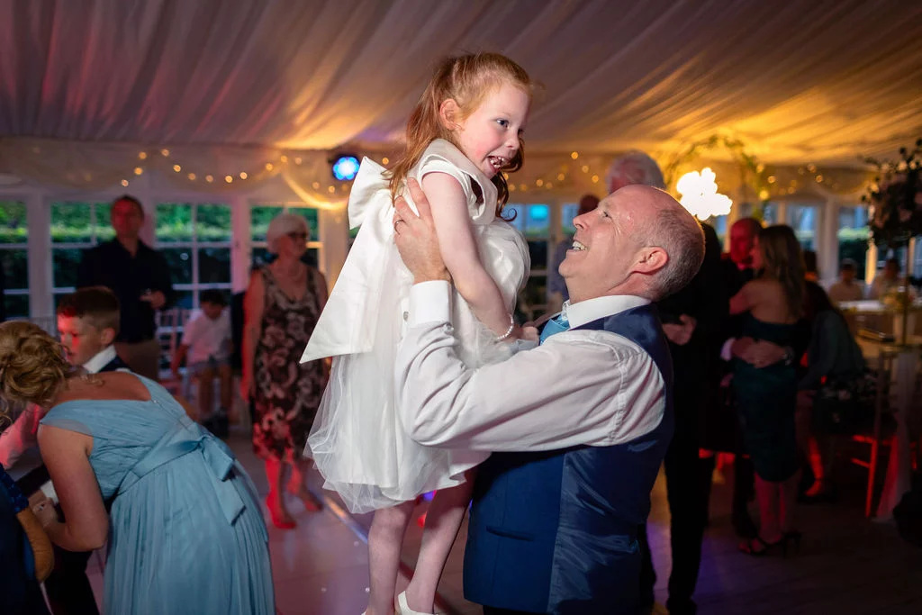 A smiling man lifts a delighted young girl in a white dress high into the air on a lively dance floor inside a draped marquee.