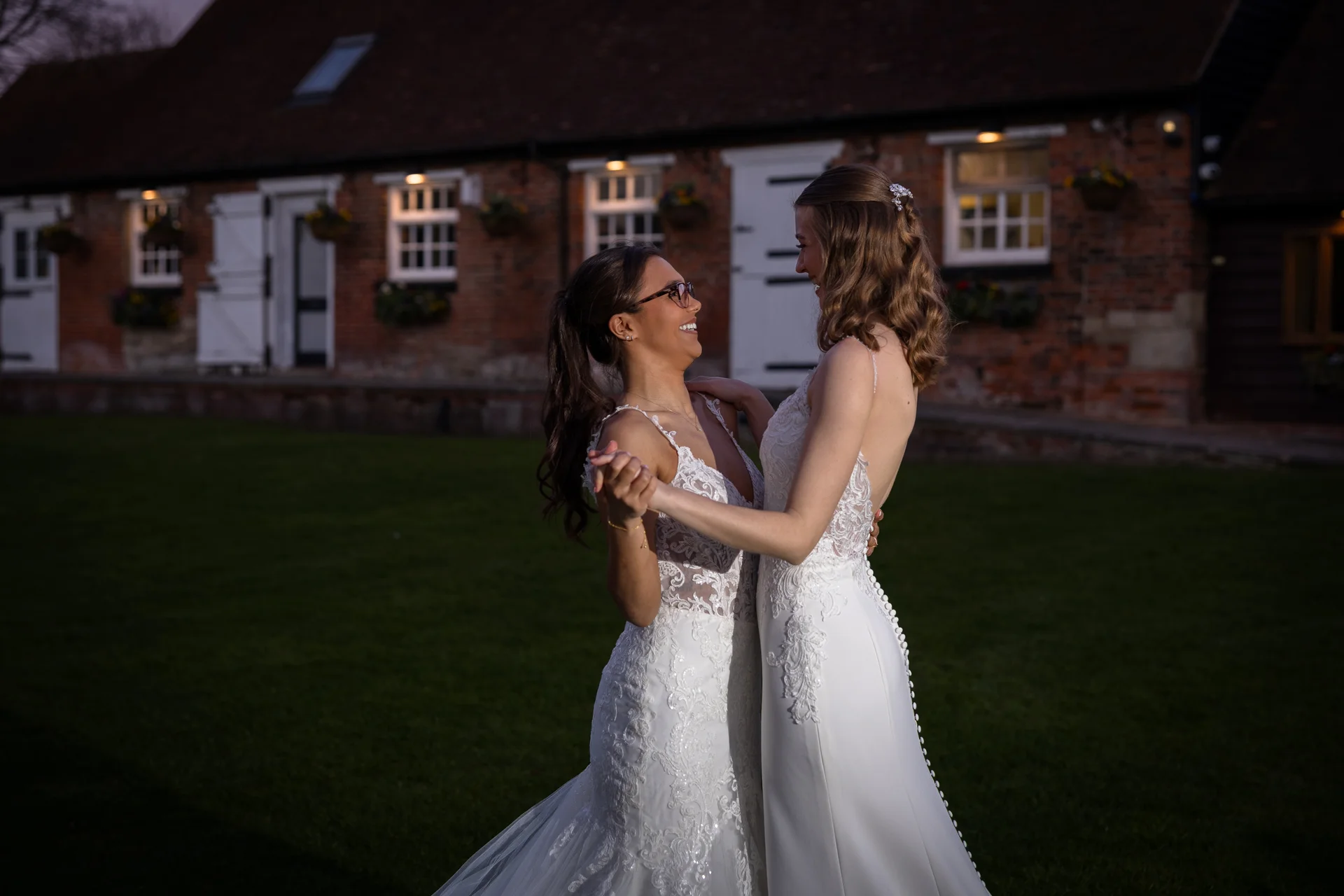 Two smiling brides in white lace and satin wedding gowns share a dance outdoors in front of a rustic brick barn at dusk.