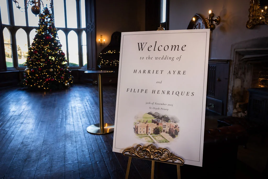 A elegant wedding welcome sign on an ornate easel stands in a historic country house hall featuring a decorated Christmas tree and large windows.