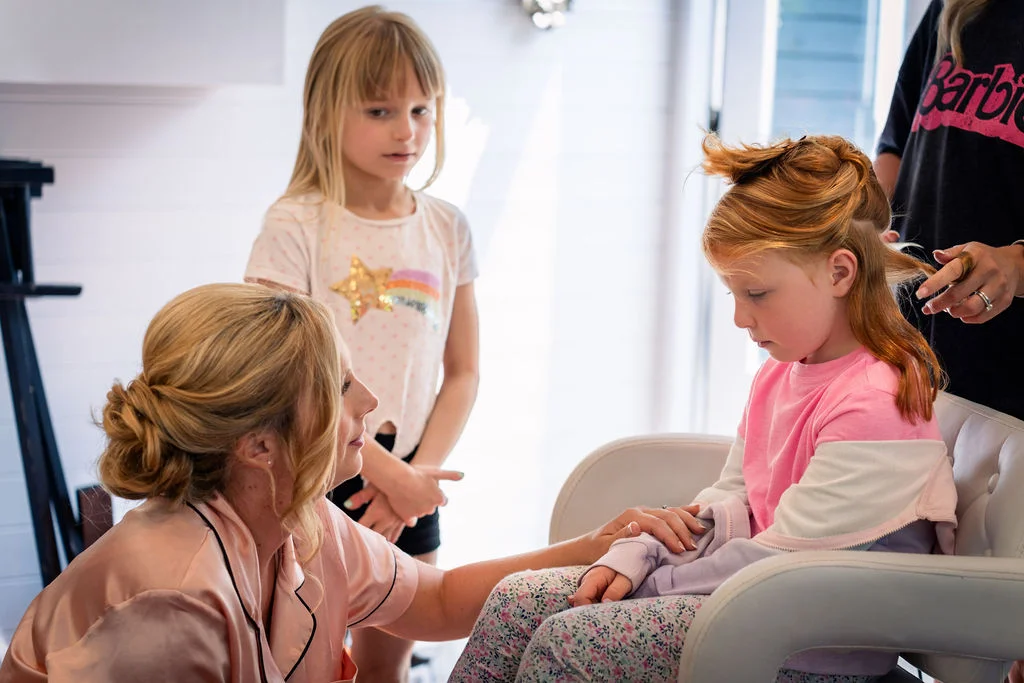 A bride in a silk robe gently holds the hand of a young flower girl sitting in a styling chair having her hair done, while another child looks on.
