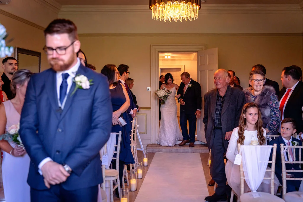 The emotional groom stands waiting at the altar in a navy suit as the bride enters the ceremony room escorted by her father.