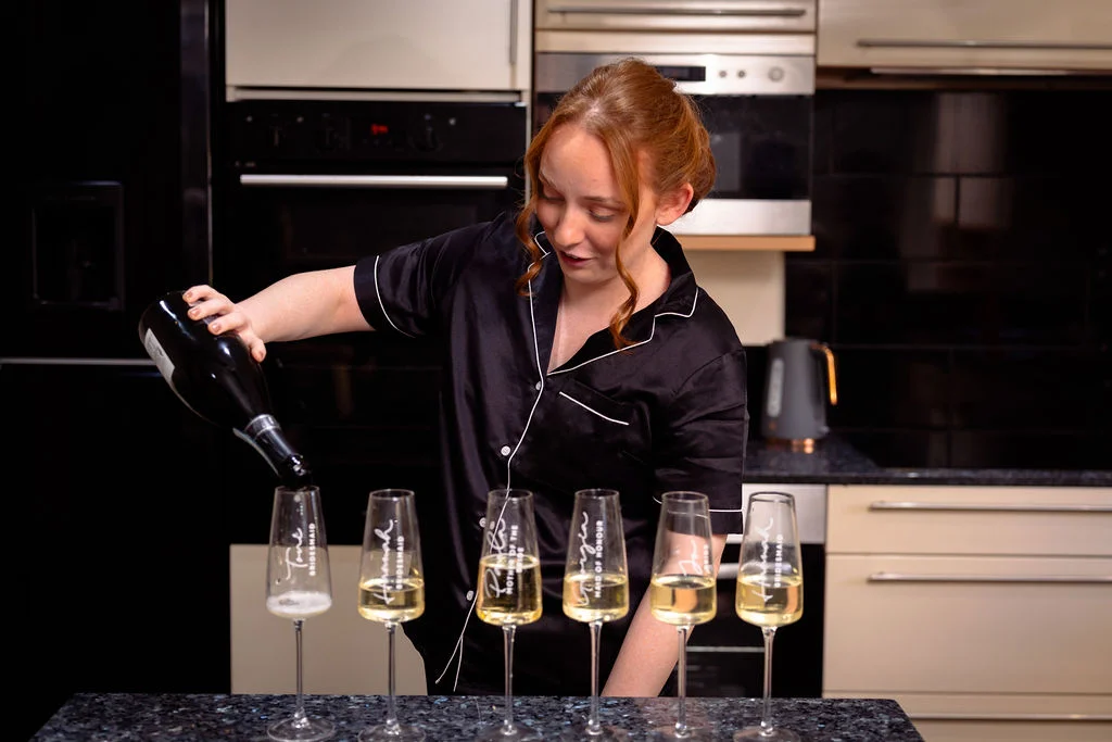 A bridesmaid in black satin pyjamas pours champagne into a row of personalised flutes in a kitchen during morning bridal preparations.
