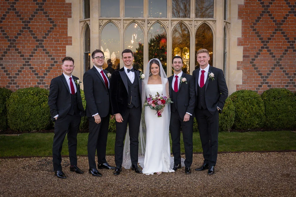 The bride and groom pose outdoors with four groomsmen in black suits and red ties in front of a grand manor house window.