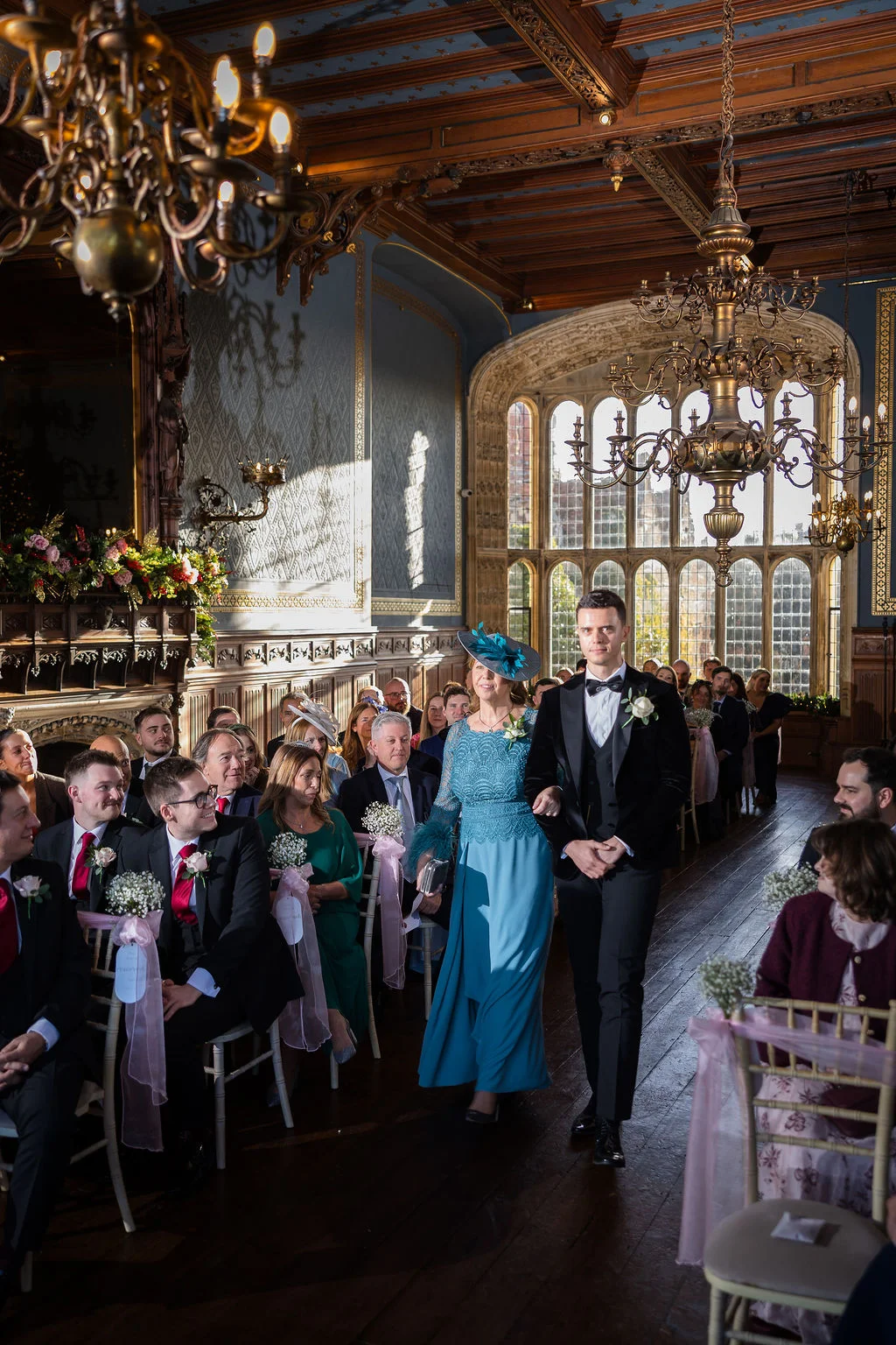 A groom and mother of the bride walk down the aisle together in a grand panelled hall with large arched windows and elaborate chandeliers during a wedding ceremony.