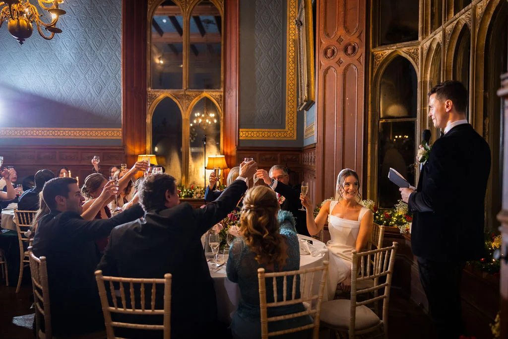 A groom delivers a speech at a grand castle wedding reception while guests raise their glasses in a toast around a candlelit round table.