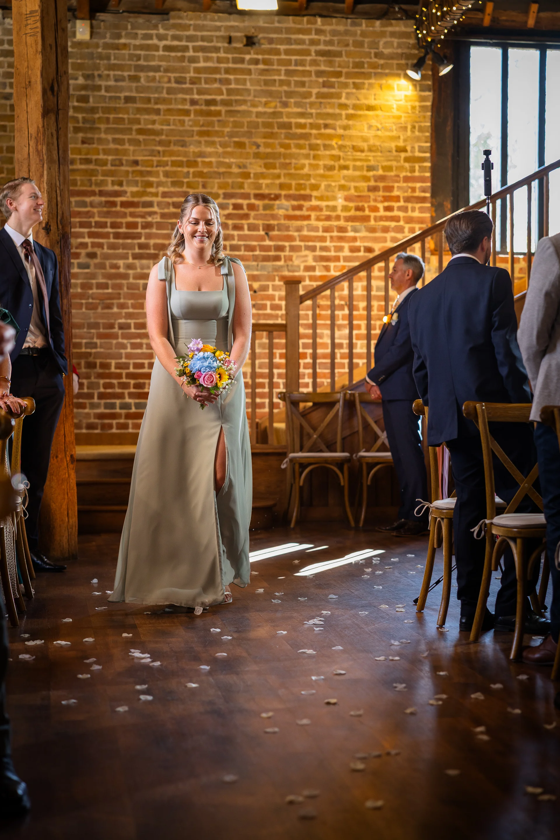 A bridesmaid walks down the wooden aisle of a rustic barn venue with exposed brick walls, holding a colourful bouquet and wearing a sage green dress with a thigh split.