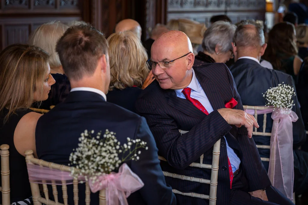 An older guest in a pinstripe suit and red tie chats with a seated guest during a wedding ceremony in a wood-panelled room.