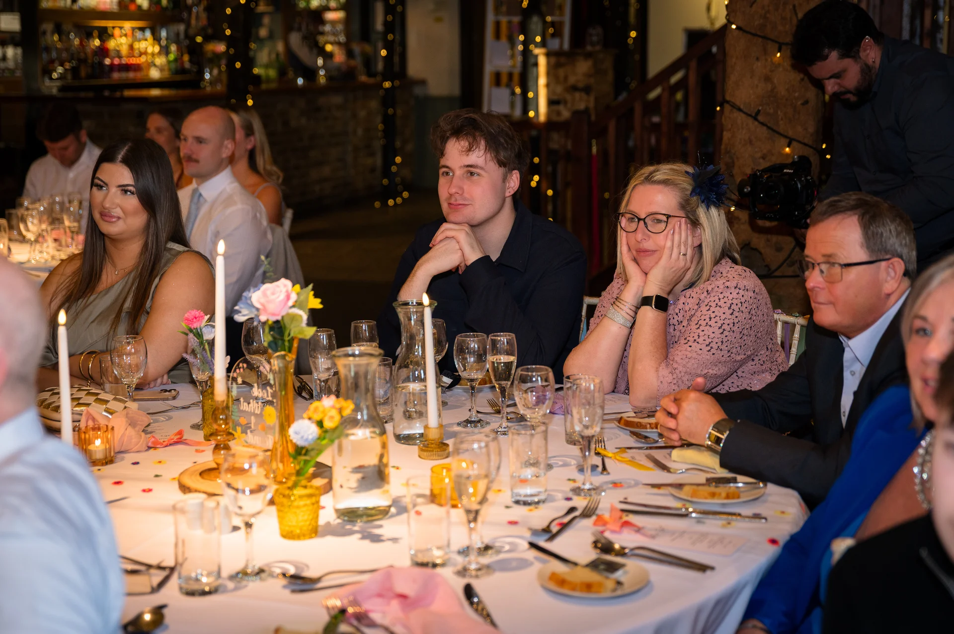 Wedding guests are seated at a round banquet table enjoying speeches during the evening reception.