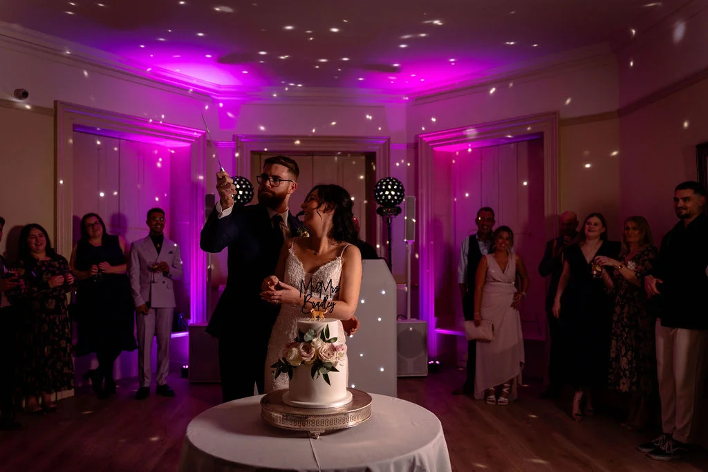 A smiling bride and groom cut into a white two-tiered wedding cake beneath vibrant pink lighting and shimmering disco ball reflections while guests watch.