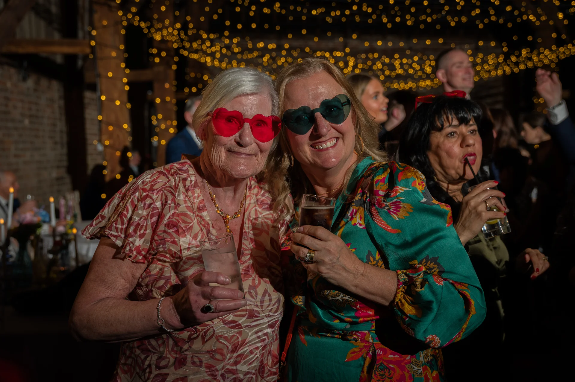 Two smiling guests wearing novelty heart-shaped sunglasses hold drinks at a lively evening wedding reception under a canopy of fairy lights inside a rustic barn.