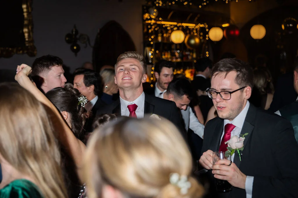 Guests and groomsmen enjoy dancing and drinks on the lively evening reception dance floor beneath warm string lighting.