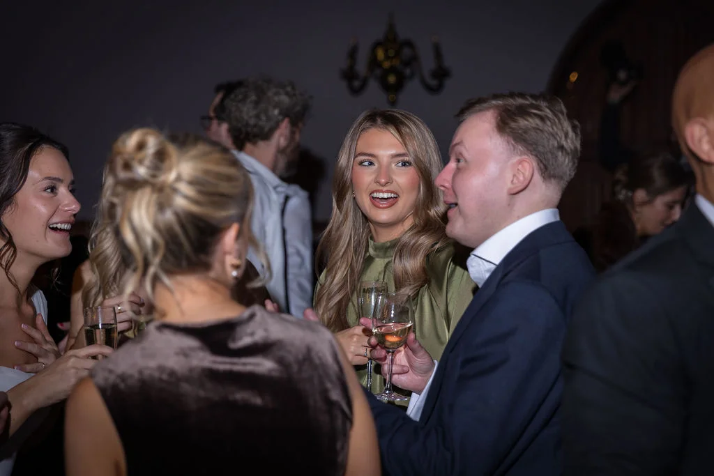 Wedding guests laugh and chat animatedly while holding glasses of wine during an evening drinks reception.