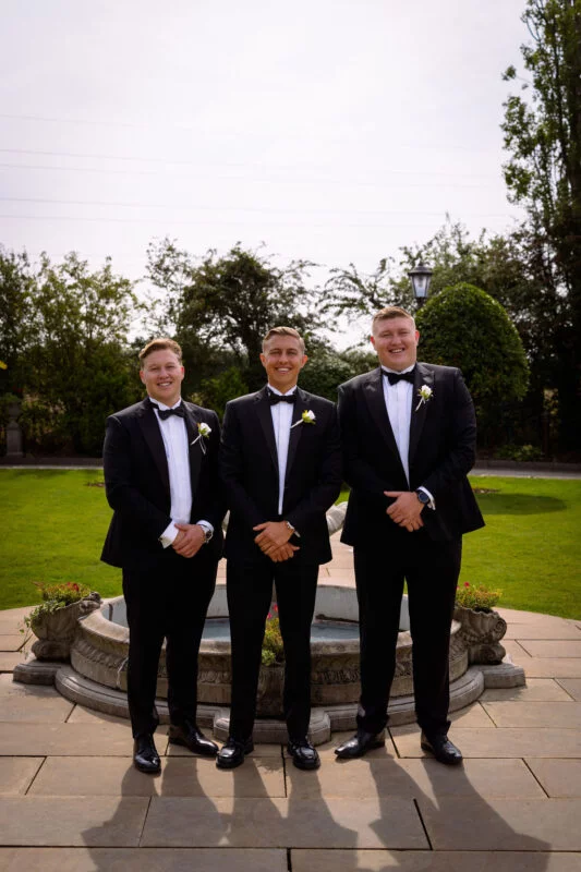 Three smiling groomsmen in matching black tuxedos and bow ties pose together outdoors in front of a stone courtyard fountain.