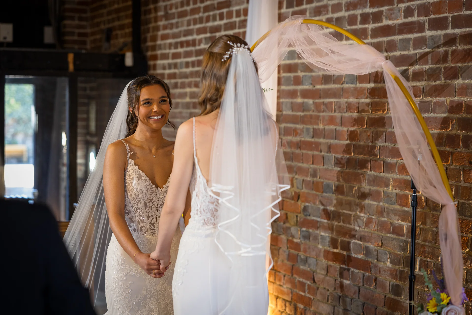 Two smiling brides holding hands during their wedding ceremony before an exposed brick wall and a decorative pink-draped arch.