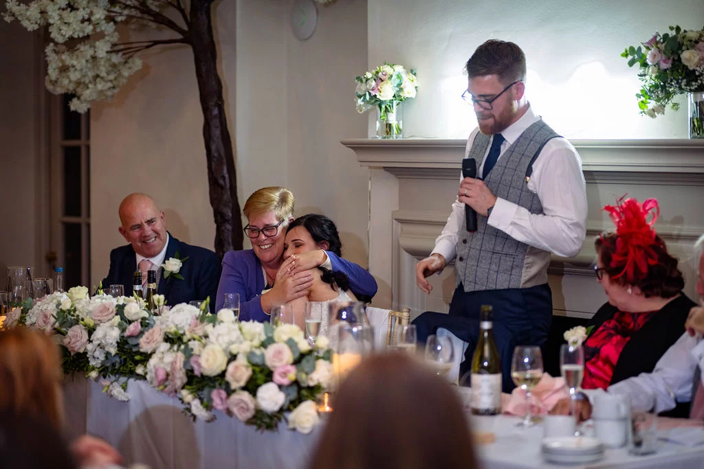 A groom delivers a speech at the top table while guests laugh and embrace during the wedding reception.