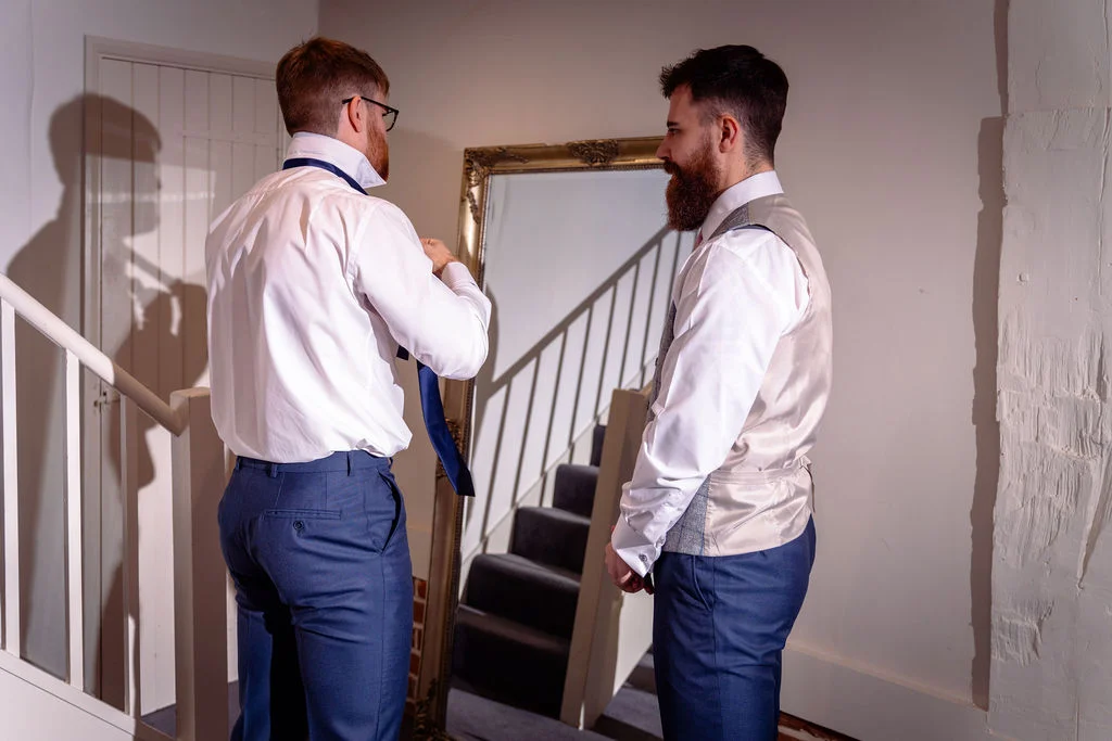 Two grooms getting ready together, with one adjusting his tie in front of a mirror reflecting a staircase.